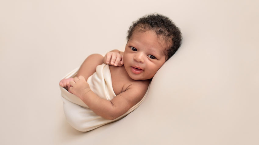 Gainesville newborn photos baby posed on his back on neutral cream blanket big brown baby eyes wide open baby Chad grinning at camera head full of curly brown bsby hair