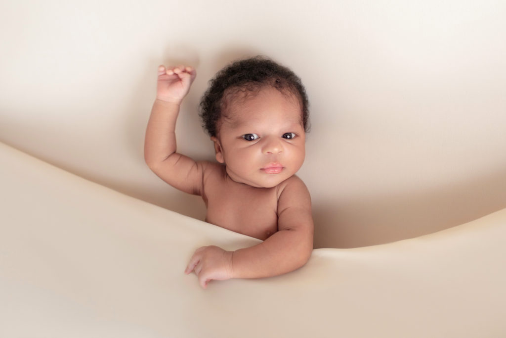 Gainesville newborn photos baby posed on his back on neutral cream blanket big brown eyes wide open arm up waving head full of curly brown hair