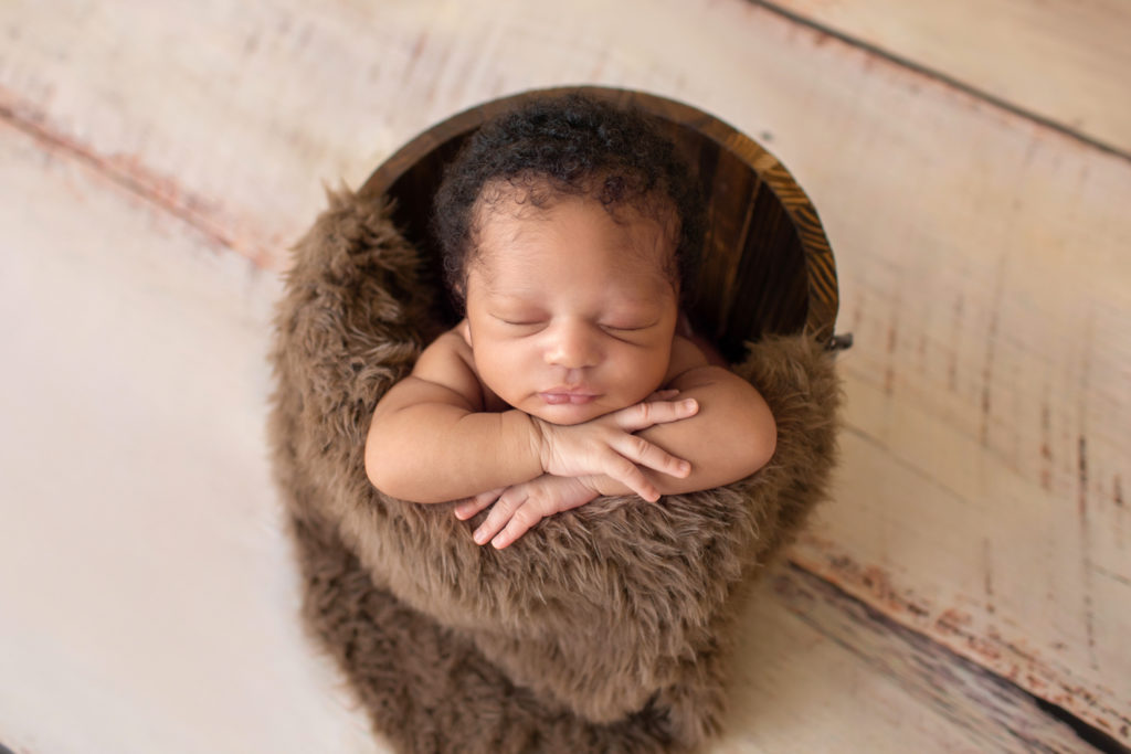 Gainesville Newborn Photos baby sleeping in a brown fur stuffed wooden bucket with head propped on his little hands