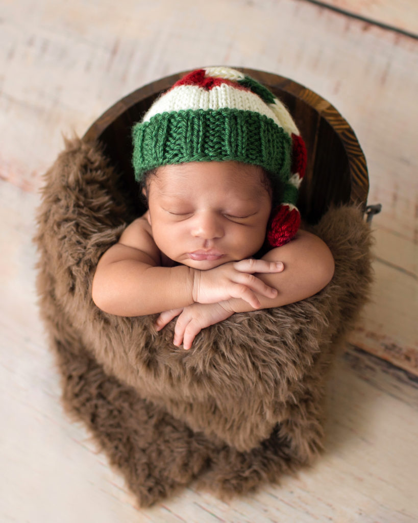 Gainesville Newborn Photos baby sleeping in a brown fur stuffed wooden bucket with head propped on his little hands Christmas sleepy hat on his head