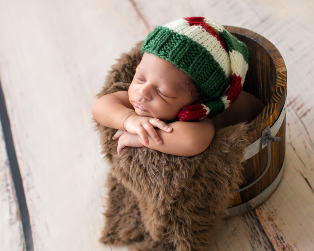 Gainesville Newborn Photos baby sleeping in a brown fur stuffed wooden bucket with head propped on his little hands Christmas sleepy hat on his head photographed with backlight