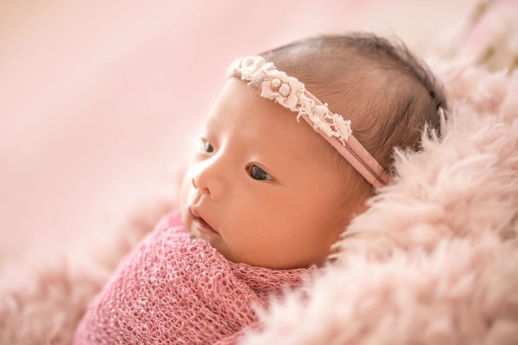 Gainesville newborn portraits baby girl with her eyes open wrapped in dusty pink fabric and lying on pale pink fur with flowers photo shot with backlight