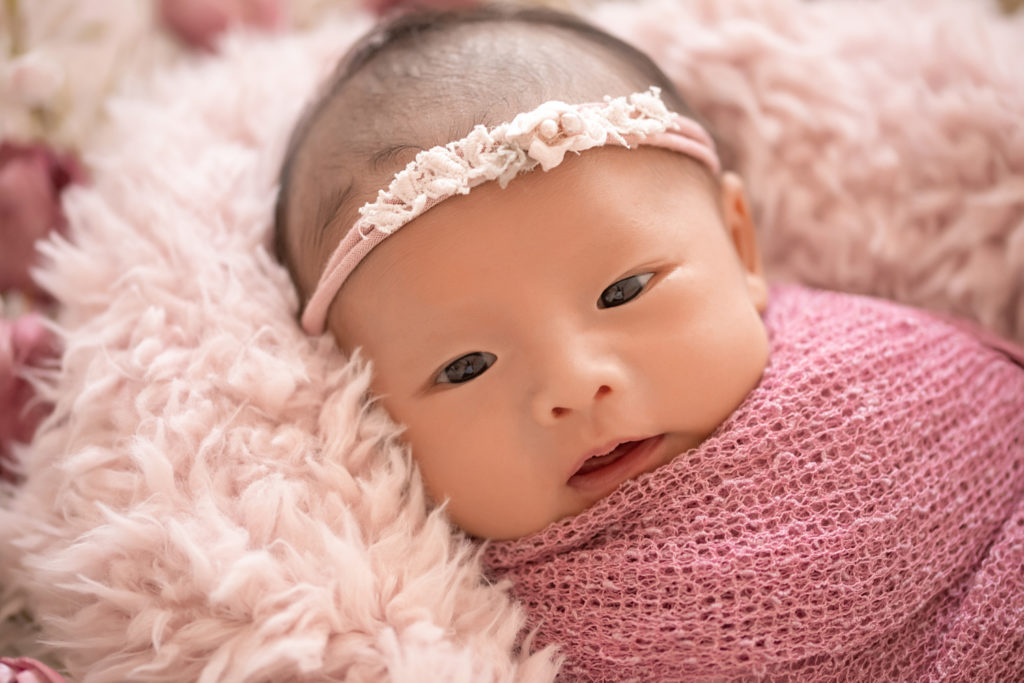 Gainesville newborn portraits baby girl looking straight at camera with her eyes open wrapped in dusty pink fabric and lying on pale pink fur with flowers