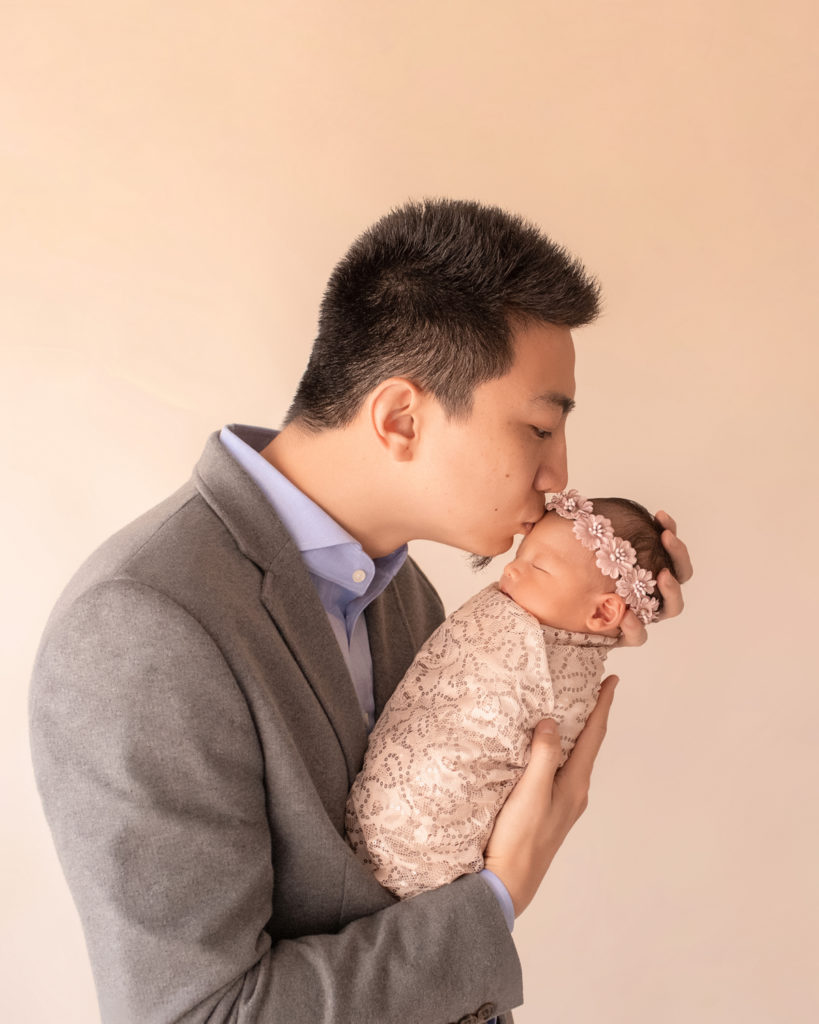 Gainesville newborn portraits profile of dad Bing giving a kiss and holding close his baby girl wrapped in cream fabric with sequins and wearing floral dusty pink crown