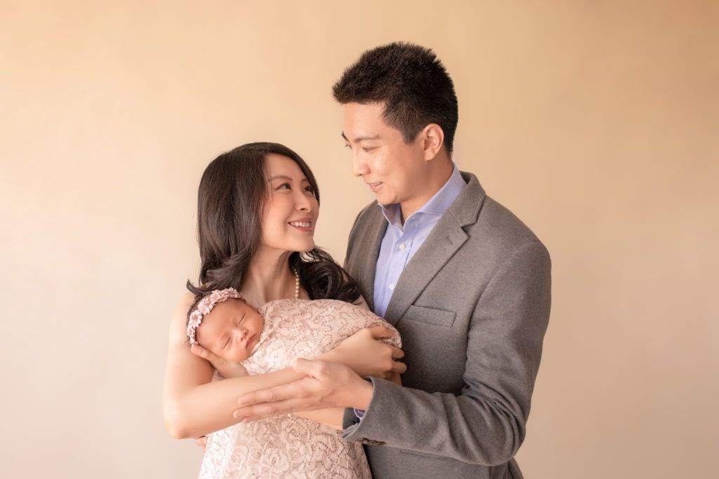 Gainesville newborn portraits gorgeous mom Vera and handsome dad Bing looking at each other and holding close their newborn baby girl wrapped in matching cream fabric with sequins and wearing floral dusty pink crown