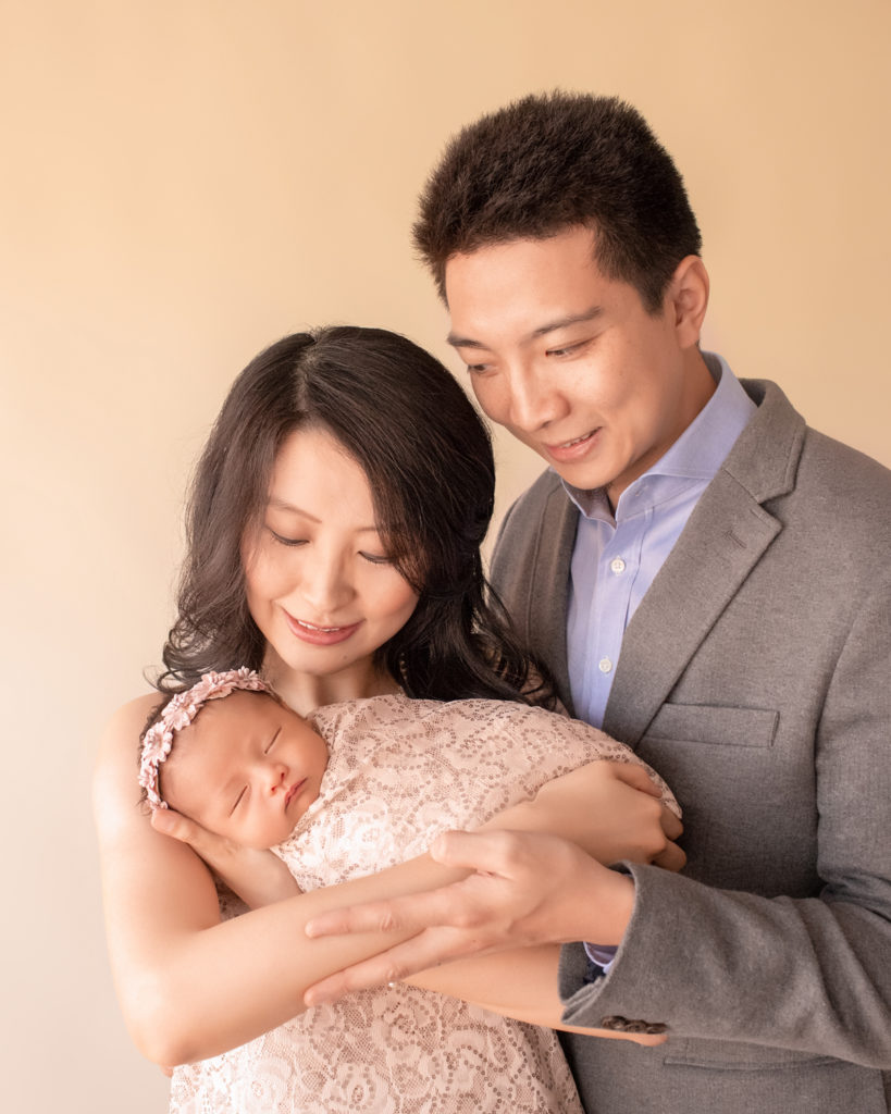 Gainesville newborn portraits beautiful mom Vera and handsome dad Bing holding close their baby girl wrapped in matching cream fabric with sequins and wearing floral dusty pink crown