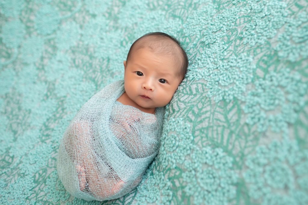 Gainesville newborn portraits baby girl looking straight at camera with her eyes open wrapped in aqua fabric and lying on pale aqua floral blanket