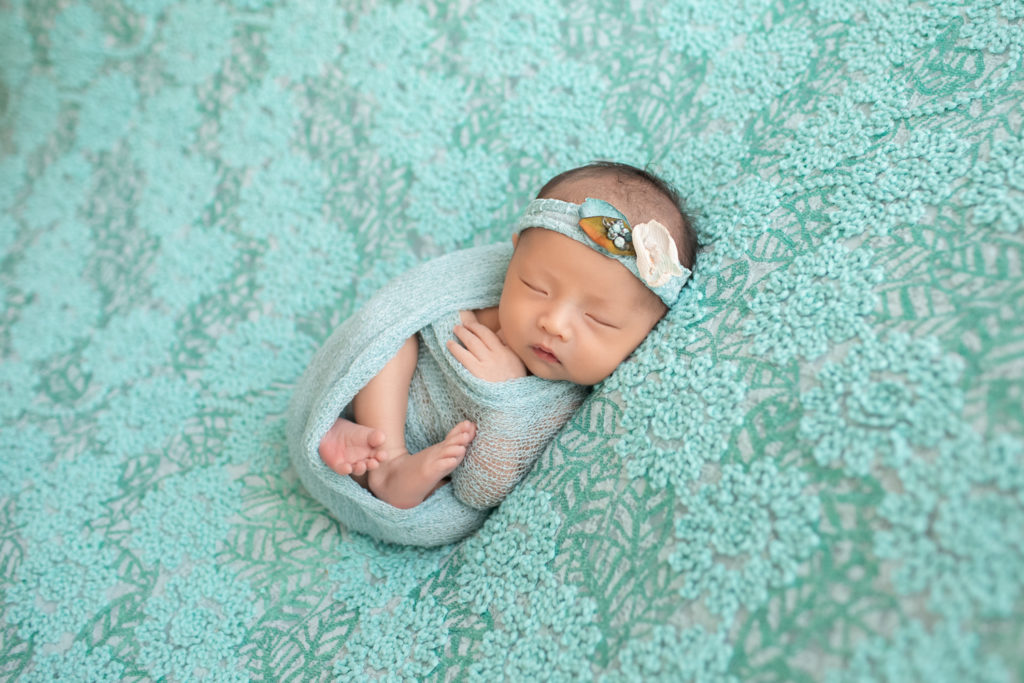 Gainesville newborn portraits baby girl with aqua floral head tie wrapped in matching aqua fabric and lying on pale aqua floral blanket hand close to face