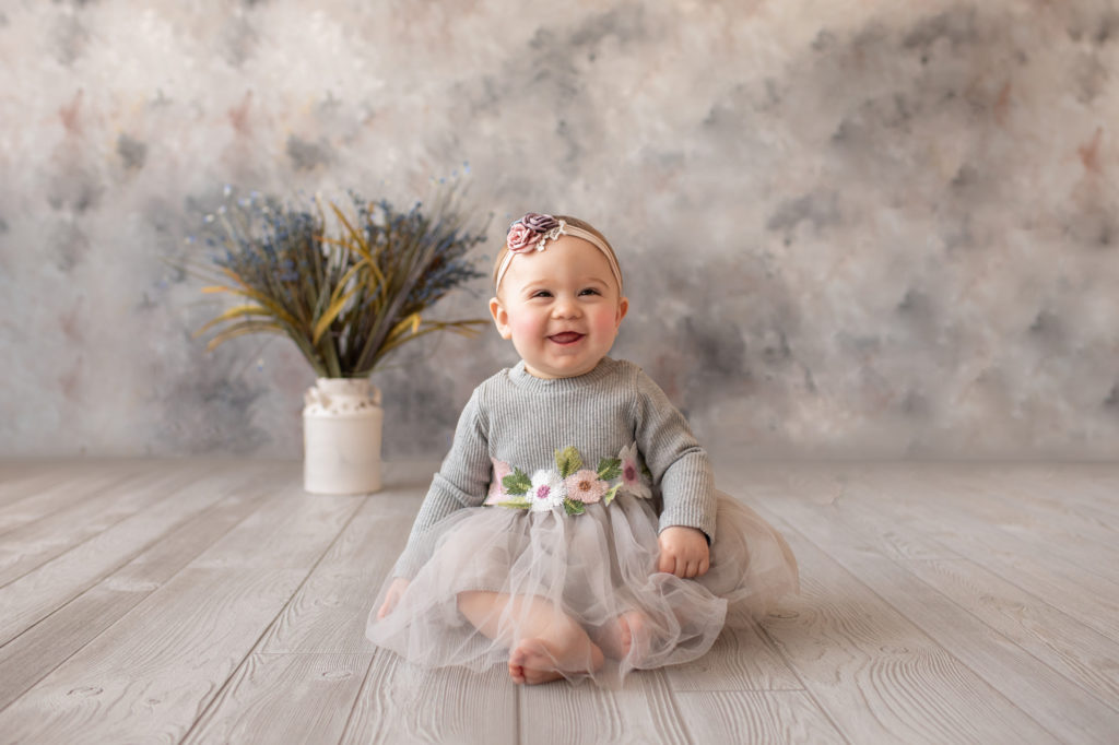 baby photo eight month girl Madison dressed in grey floral dress and pink floral headband with the biggest baby grin sitting by herself on grey floor with floral backdrop Gainesville Florida