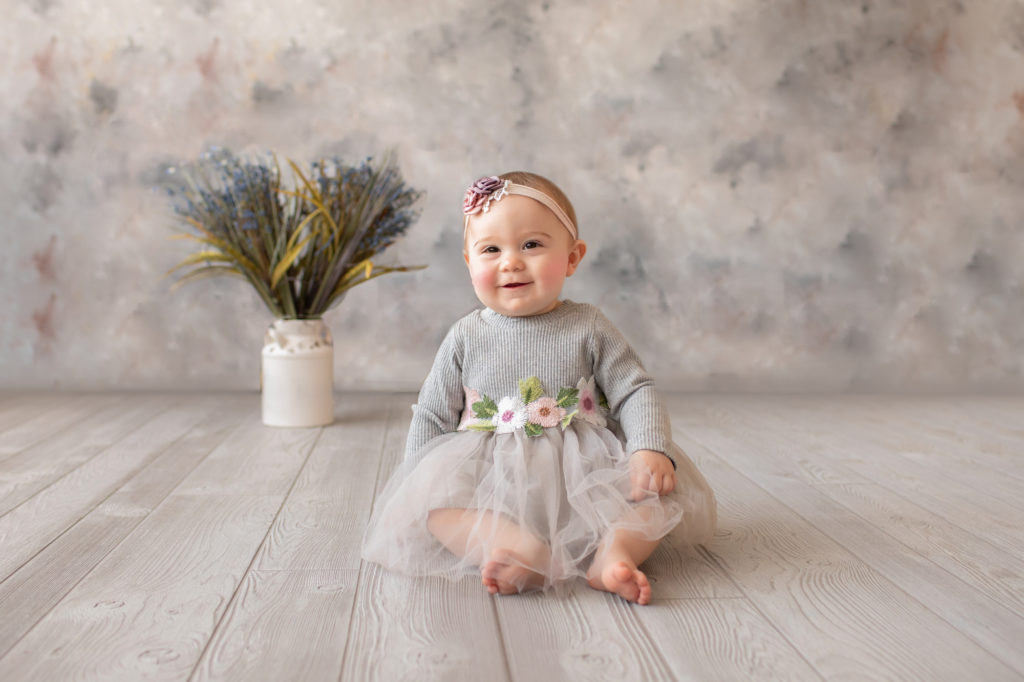 baby photo eight month girl Madison dressed in grey floral dress and pink floral headband smiling and sitting by herself on grey floor with floral backdrop Gainesville Florida