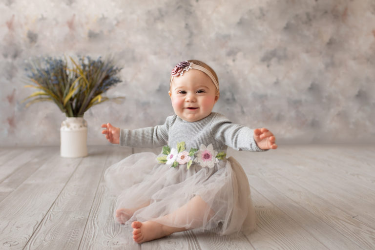 baby photo eight month girl Madison dressed in grey floral dress and pink floral headband with arms out to side sitting by herself on grey floor with floral backdrop Gainesville Florida