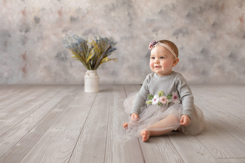 baby photo eight month girl Madison dressed in grey floral dress and pink floral headband sitting by herself looking at her mom on grey floor with floral backdrop Gainesville Florida