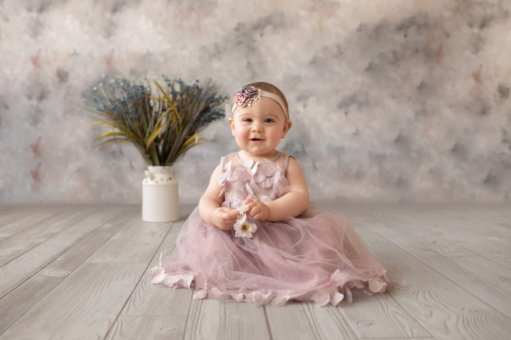 baby photos eight month girl Madison with her best smile dressed in dusty pink floral dress and pink floral headband sitting by herself on grey floor with floral backdrop Gainesville Florida