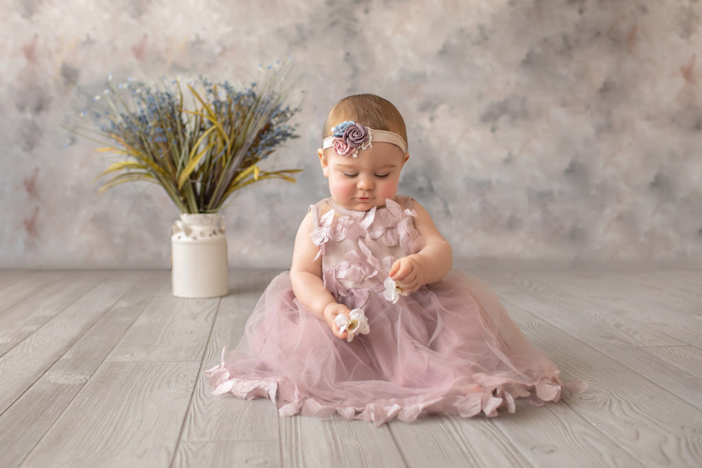 baby photo eight month girl Madison dressed in dusty pink floral dress and pink floral headband sitting by herself on grey floor with floral backdrop playing with flowers in her hands Gainesville Florida