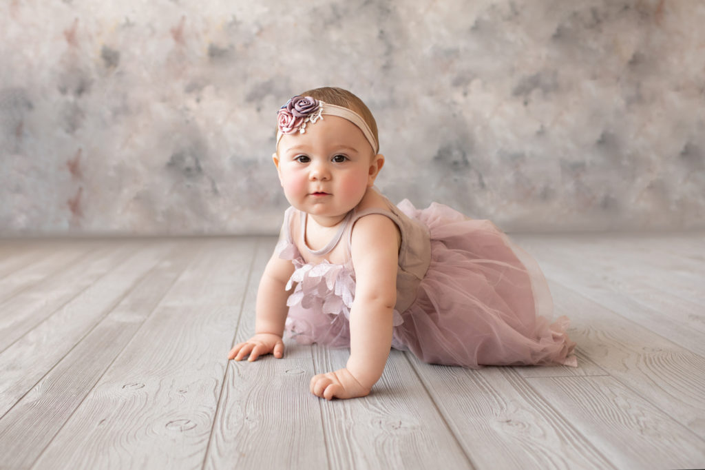 baby photo eight month girl Madison dressed in dusty pink floral dress and pink floral headband crawling on all fours on grey floor with floral backdrop Gainesville Florida