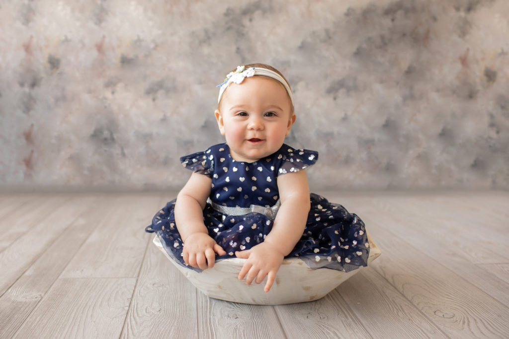 baby photo eight month girl Madison dressed in navy dress with white hearts and white floral headband sitting by herself in white bowl on grey floor with floral backdrop Gainesville Florida