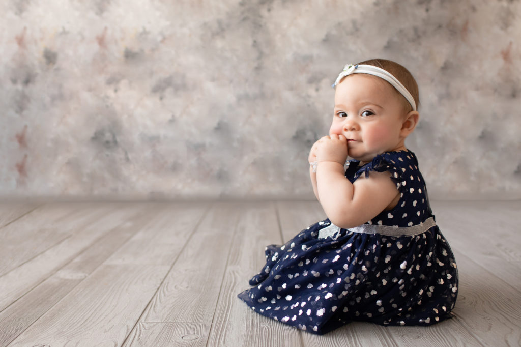 baby photo eight month girl Madison dressed in navy dress with white hearts and white floral headband sitting by herself in profile on grey floor with floral backdrop Gainesville Florida