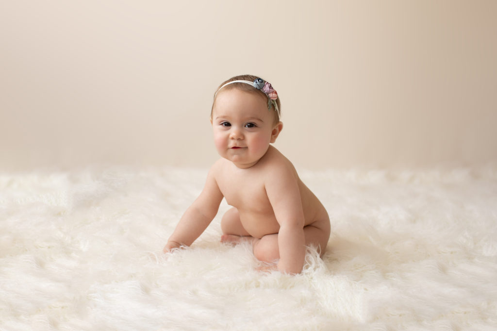 baby photo eight month girl Madison naked soft baby skin pink floral headband sitting by herself in profile on white fur with cream backdrop Gainesville Florida