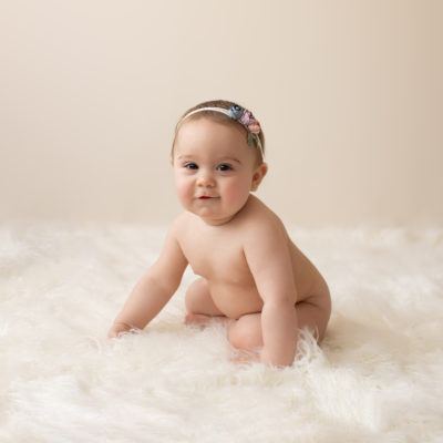 eight month girl Madison naked baby photos soft baby skin pink floral headband sitting by herself in profile on white fur with cream backdrop Gainesville Florida
