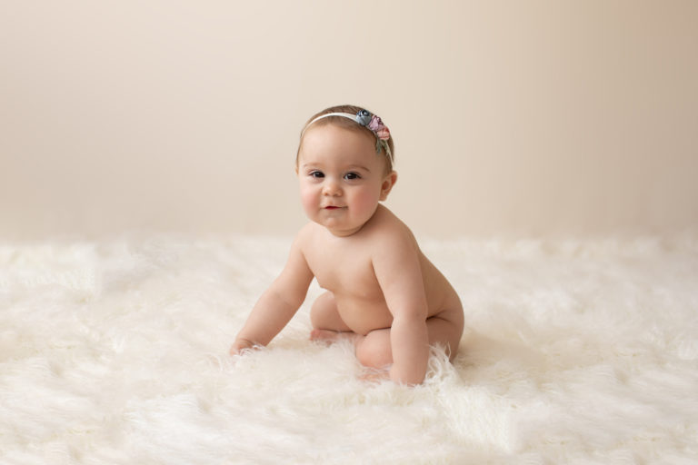 eight month girl Madison naked baby photos soft baby skin pink floral headband sitting by herself in profile on white fur with cream backdrop Gainesville Florida