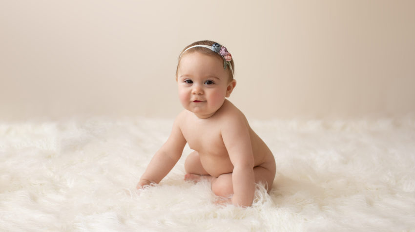 eight month girl Madison naked baby photos soft baby skin pink floral headband sitting by herself in profile on white fur with cream backdrop Gainesville Florida