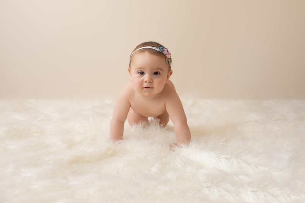 baby photos eight month girl Madison naked soft baby skin pink floral head looking at camera and crawling on white fur with cream backdrop Gainesville Florida