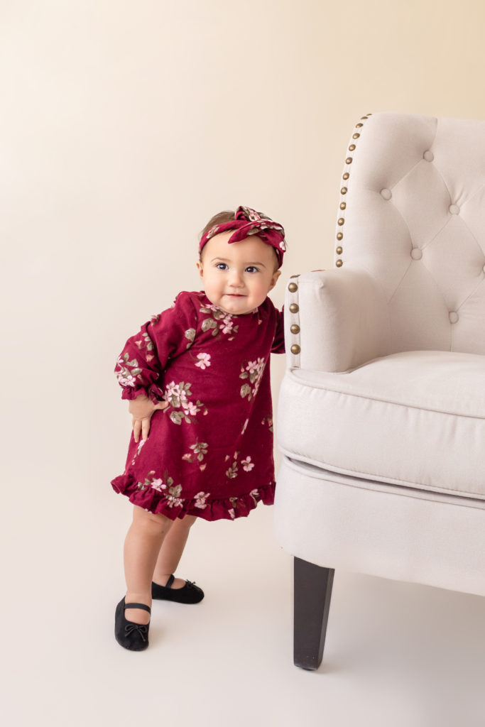 One year twin baby photo girl dressed in burgundy floral dress and headband standing by herself holding onto chair Gainesville Florida