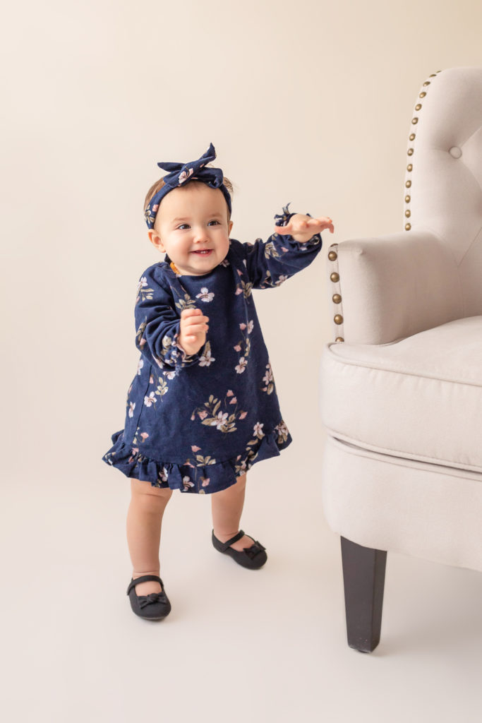 One year twin baby photo girl dressed in navy floral dress and headband standing by herself almost walking first baby steps Gainesville Florida