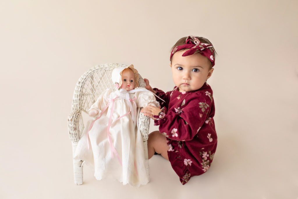 One year twin baby photo girl dressed in burgundy floral dress and headband sitting on cream floor playing with baby doll in wicker chair Gainesville Florida