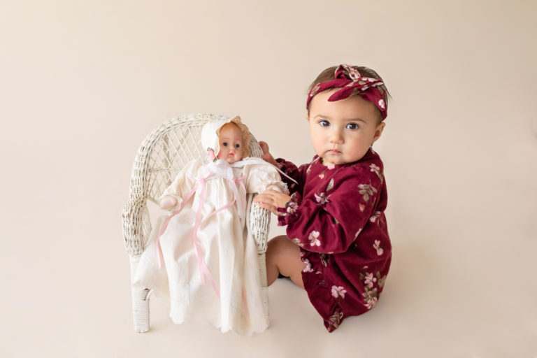 One year twin baby photo girl dressed in burgundy floral dress and headband sitting on cream floor playing with baby doll in wicker chair Gainesville Florida