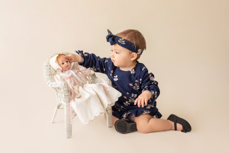 One year twin baby photo girl dressed in navy floral dress and headband sitting on cream floor playing with baby doll in wicker chair Gainesville Florida
