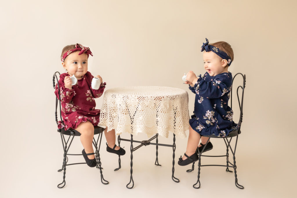 One year twin baby photo girls dressed in burgundy and navy floral dresses and headbands sitting for tea party at lace covered table Gainesville Florida