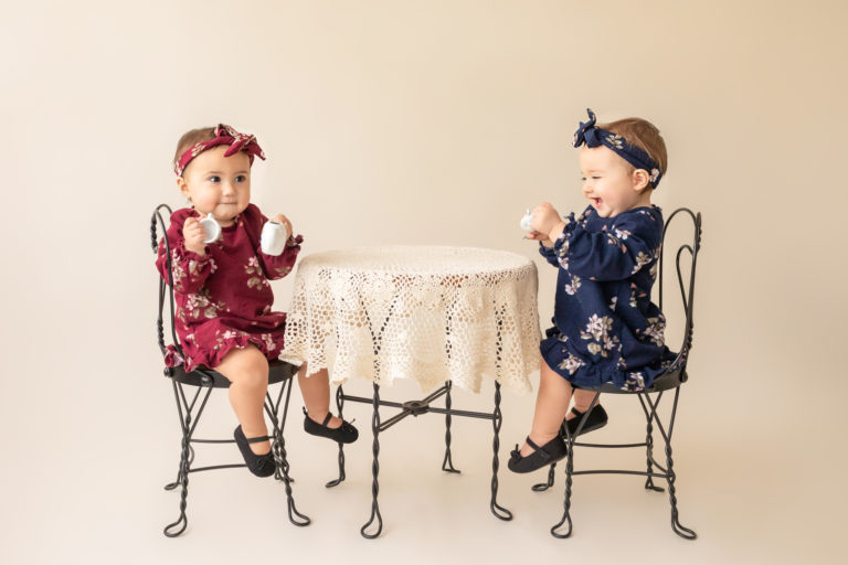 One year twin baby photo girls dressed in burgundy and navy floral dresses and headbands sitting for tea party at lace covered table Gainesville Florida