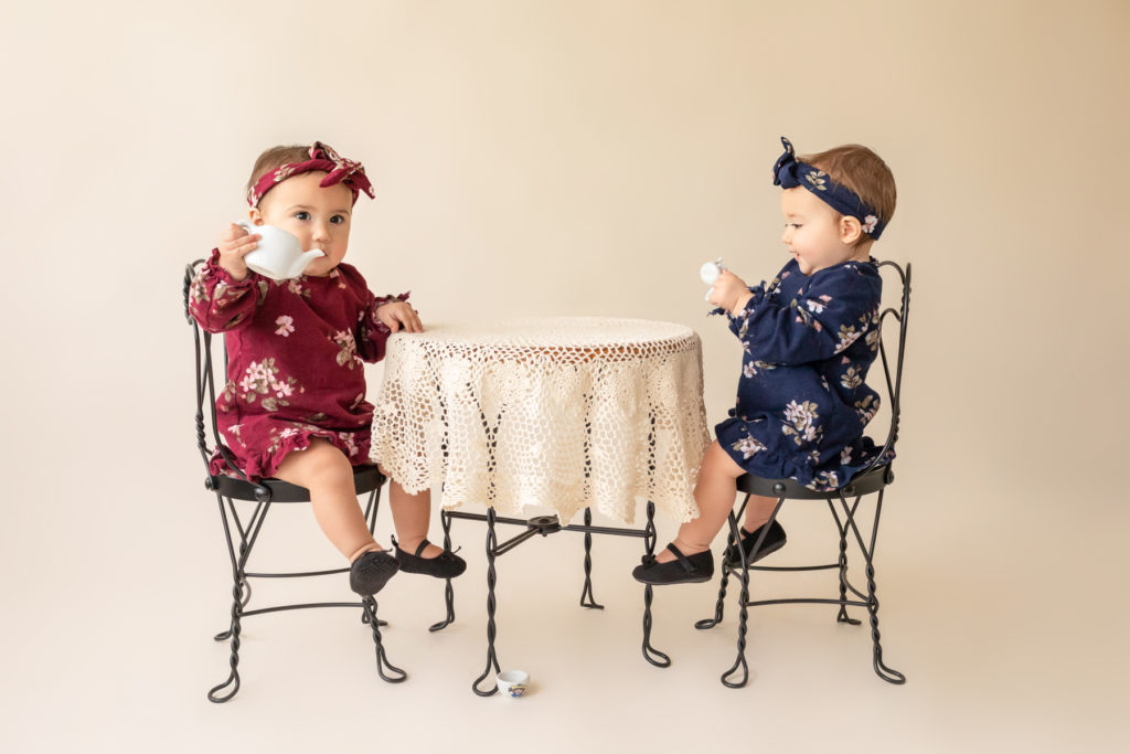 One year twin baby photo girls Renna and Winona dressed in burgundy and navy floral dresses and headbands sitting for tea party at lace covered table Gainesville Florida