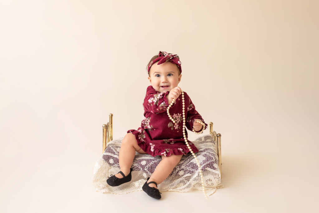 One year twin baby photo girl dressed in burgundy floral dress and headband sitting on lace covered bed playing with strand of pearls Gainesville Florida