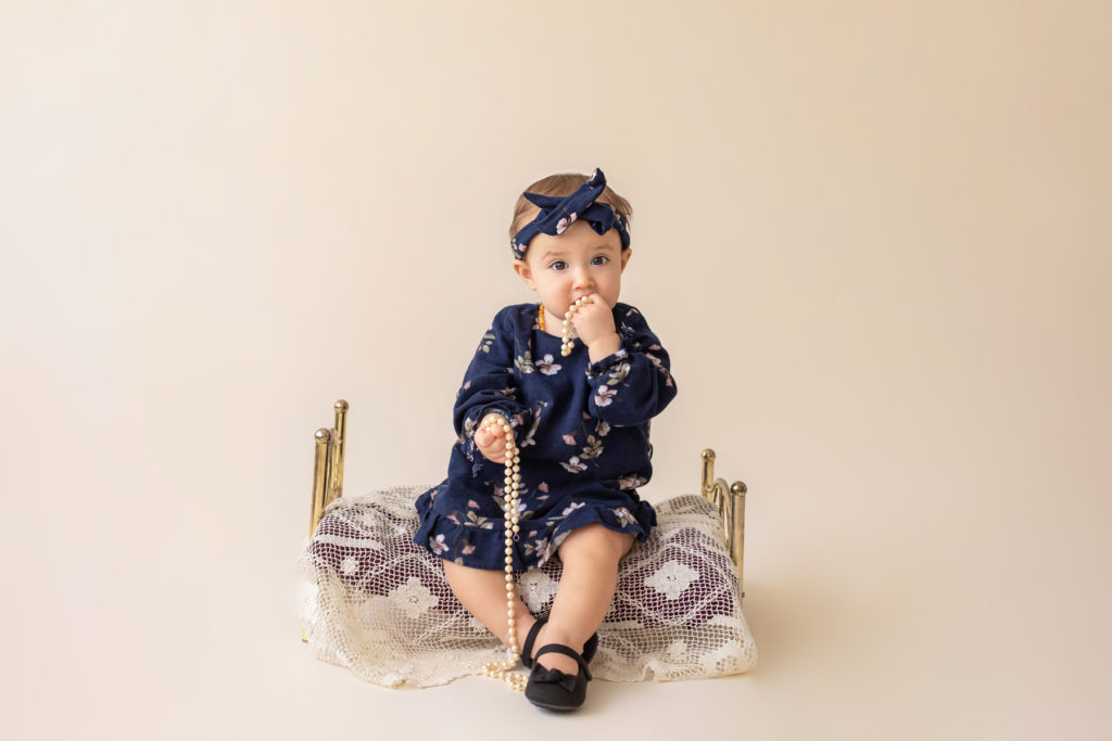 One year twin baby photo girl dressed in navy floral dress and headband sitting on lace covered bed playing with strand of pearls Gainesville Florida