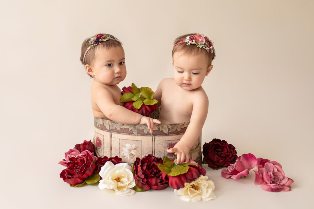 One year twin baby photo girls both naked with floral headbands sitting in same floral hat box playing with burgundy rose and ivory silk flowers Gainesville Florida