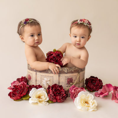 One year twin baby photo girls both naked with floral headbands sitting in same floral hat box playing with burgundy rose and ivory silk flowers looking up Gainesville Florida