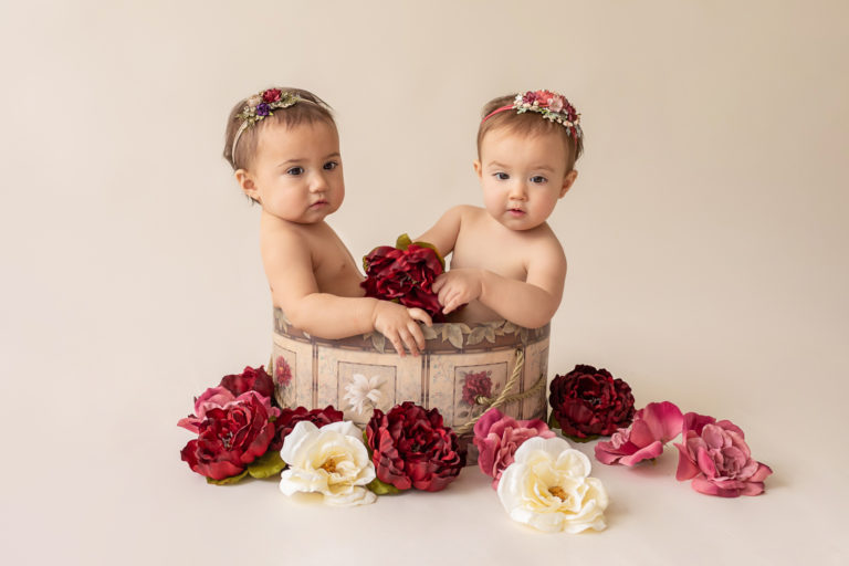 One year twin baby photo girls both naked with floral headbands sitting in same floral hat box playing with burgundy rose and ivory silk flowers looking up Gainesville Florida