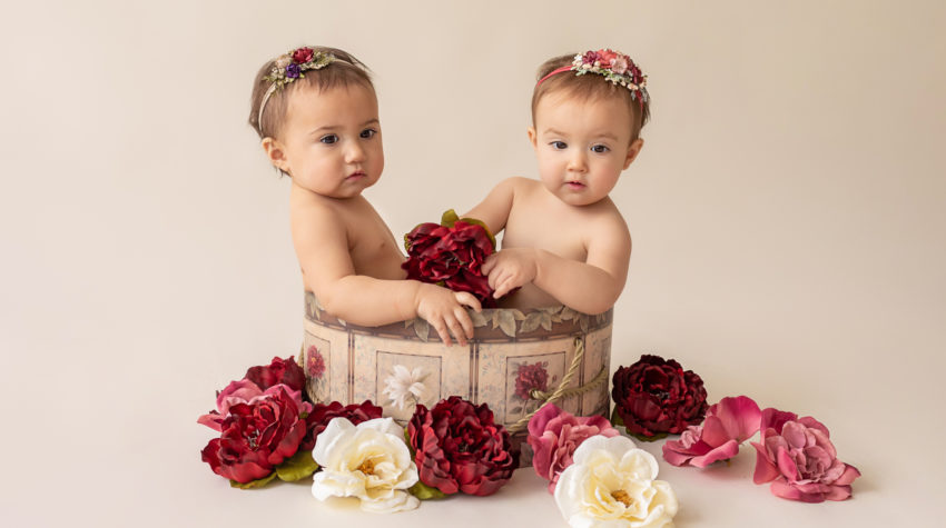 One year twin baby photo girls both naked with floral headbands sitting in same floral hat box playing with burgundy rose and ivory silk flowers looking up Gainesville Florida