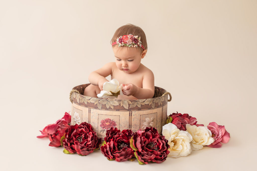 One year twin baby photo girl naked entertained with floral headband sitting in floral hat box playing with burgundy rose and ivory silk flowers Gainesville Florida