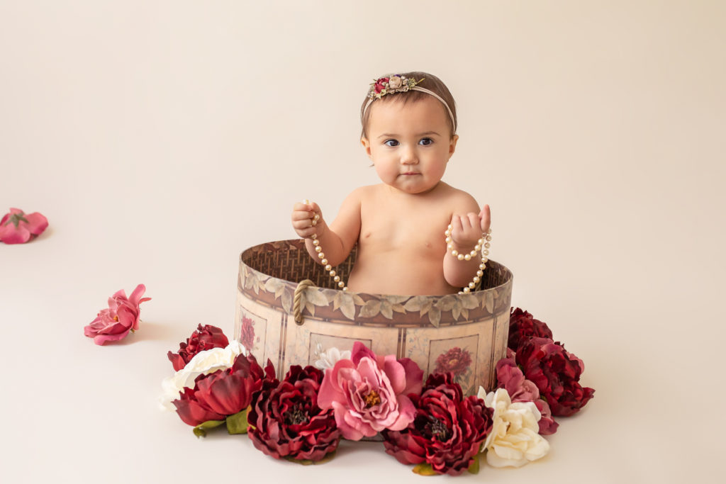 One year twin baby photo girl naked entertained with floral headband sitting in floral hat box playing entertained with strand of pearls and burgundy rose and ivory silk flowers Gainesville Florida