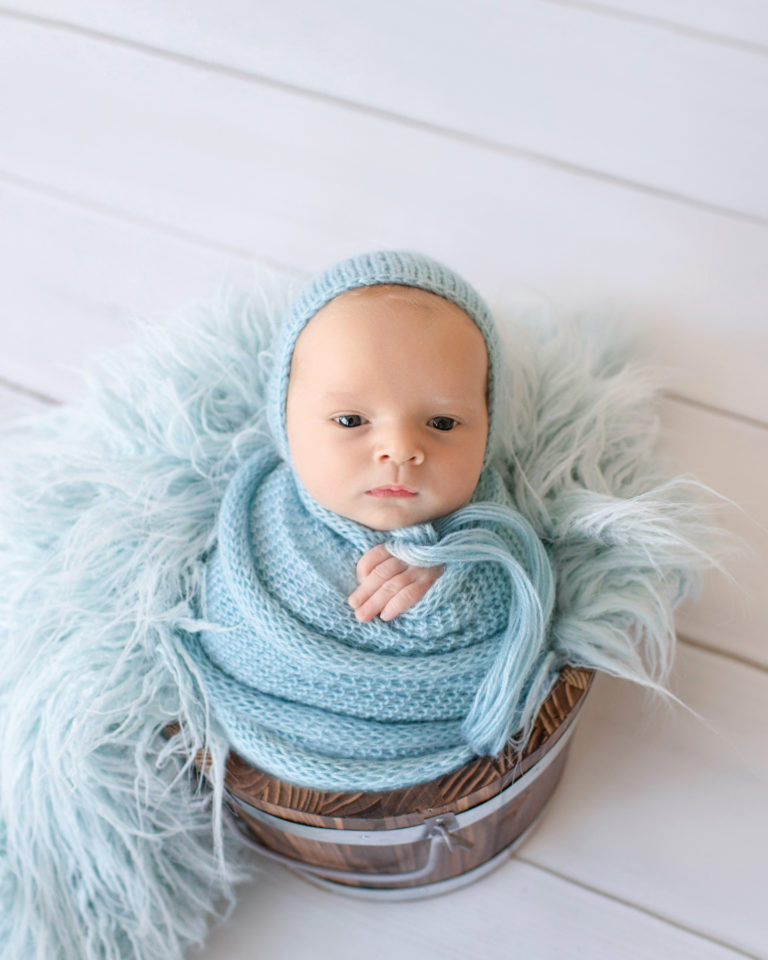 Newborn baby boy Ezra in blue wrap and blue bonnet with eyes wide open in blue fur stuffed brown bucket Gainesville Florida newborn photographer