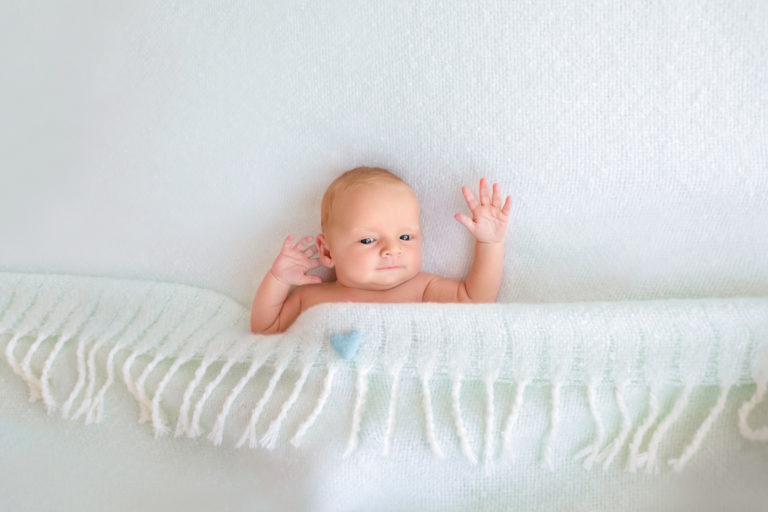 Newborn baby boy Ezra with eyes open and arms waving tucked into blanket on bed with aqua heart felted pillow Gainesville Florida newborn photographer