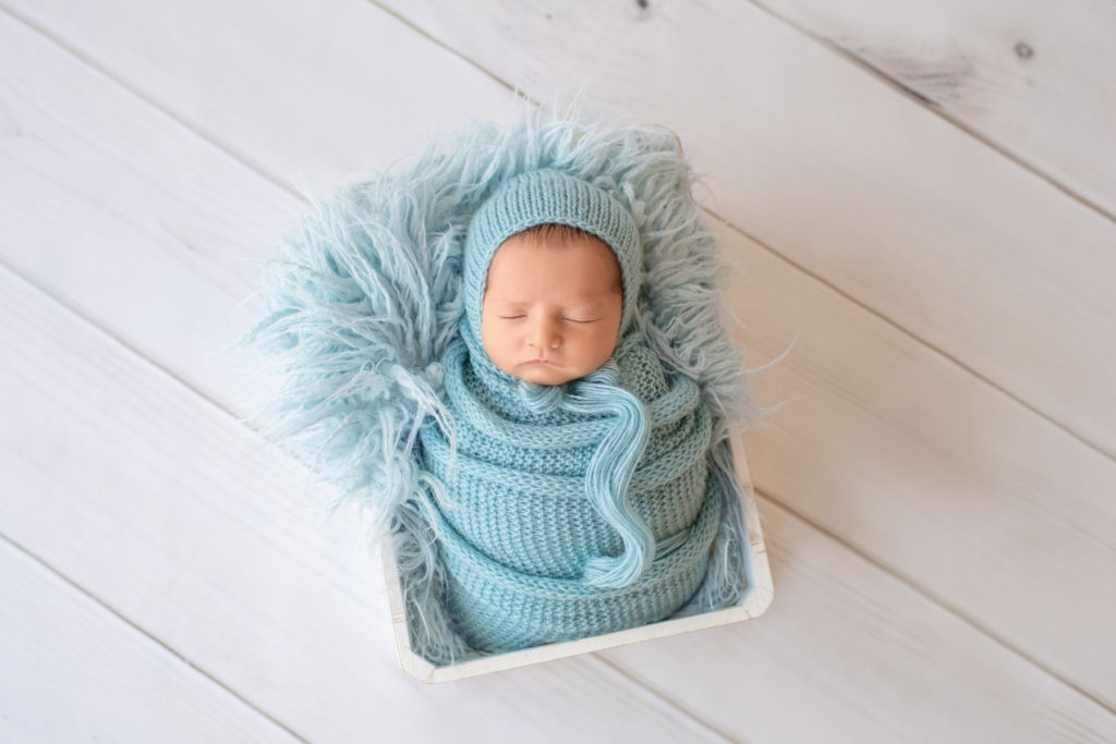 newborn Jeffery in blue handmade knit wrap and matching blue bonnet posed in blue fur stuffed white crate