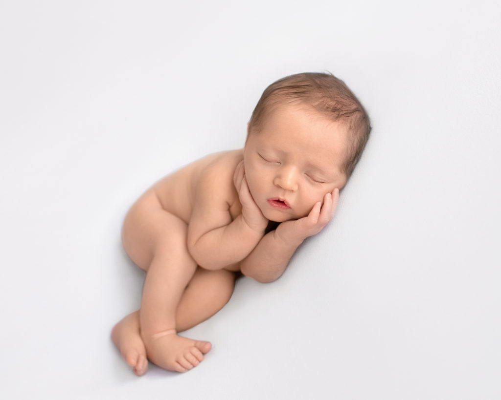 Newborn boy Jeffery with dark brown hair naked asleep with his hands wrapped around his cheeks lying on his side with his legs crossed on white blanket newborn photographer Gainesville Florida