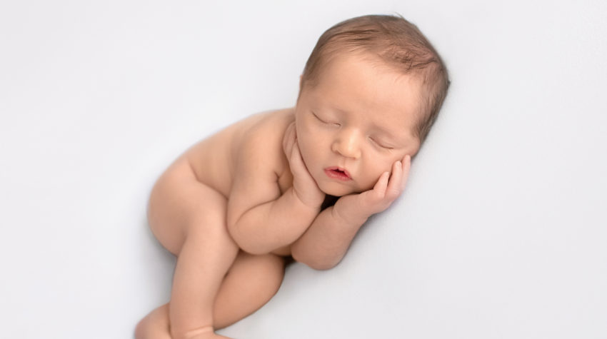 Newborn baby details boy Jeffery with dark brown hair naked asleep with his little baby hands curled around his round cheeks lying on his side with his legs crossed on white blanket newborn photographer Gainesville Florida