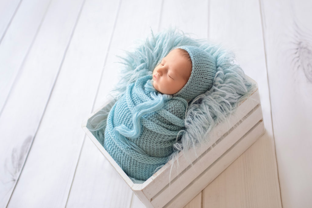 newborn Jeffery in blue handmade knit wrap and matching blue bonnet posed in blue fur stuffed white crate against backlight