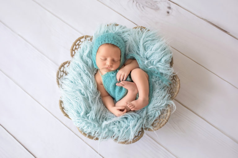 Newborn boy Jeffery dressed in handmade knitted aqua romper and matching aqua romper asleep posed on his back with arms and legs curled tight in fur stuffed brown basket newborn photographer Gainesville Florida