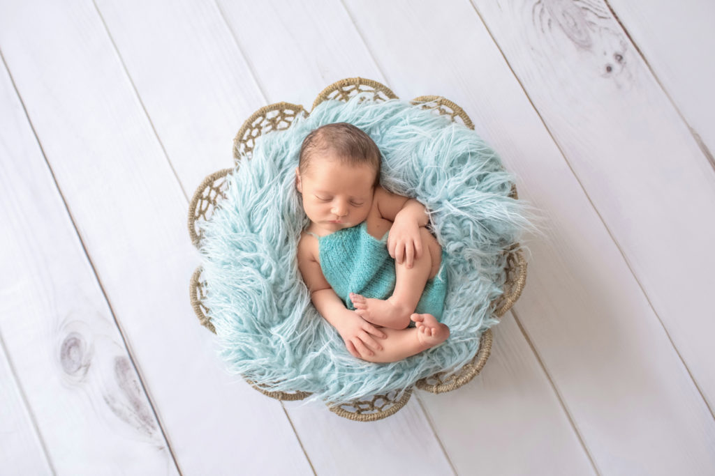 Newborn boy Jeffery dressed in aqua romper asleep posed with arms and legs curled tight on his back in fur stuffed brown basket newborn photographer Gainesville Florida